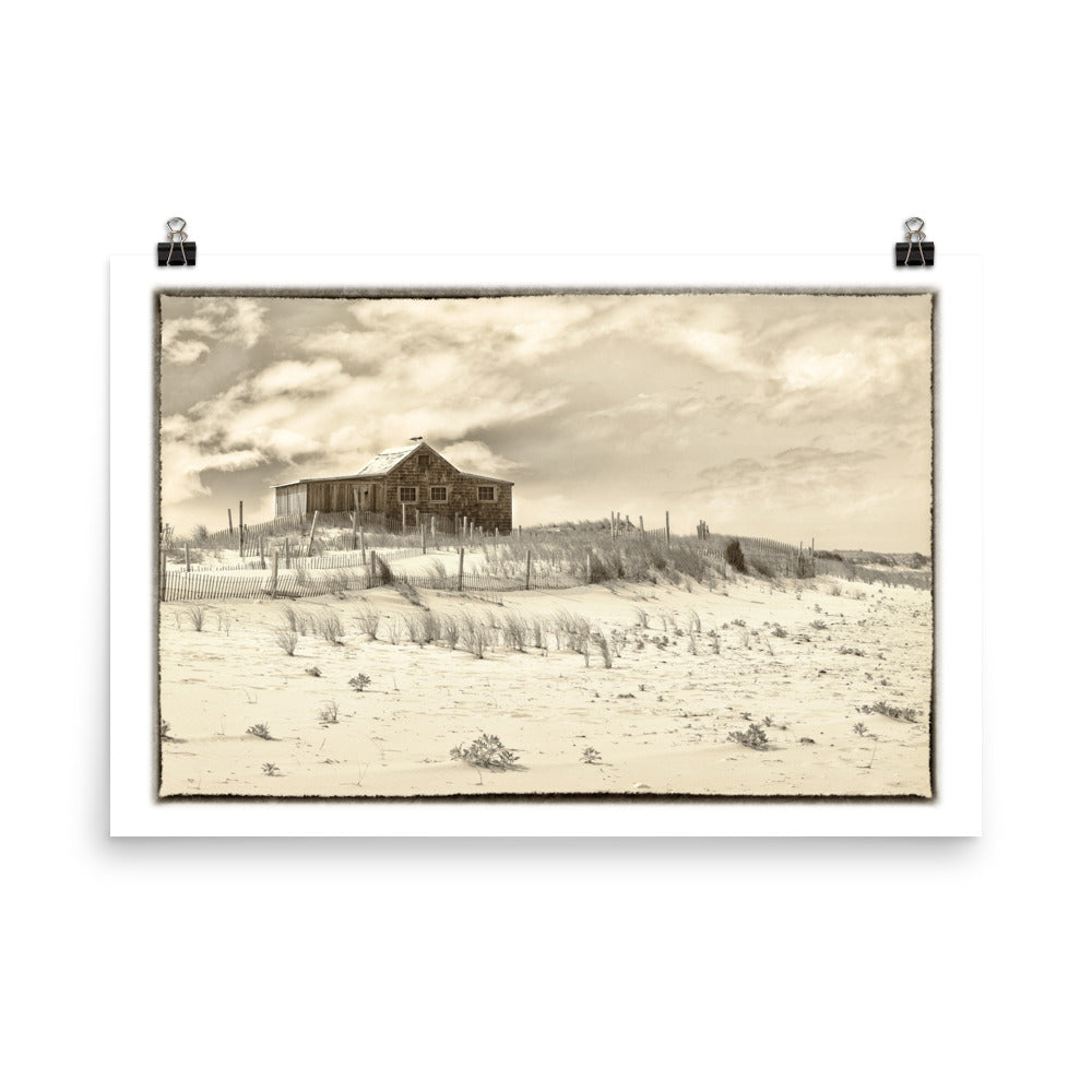 Image of The Retreat mixed media sepia tone artwork on 24 inch by 36 inch premium matte photo paper by Jessica St. Clair depicting a weathered beach cottage in the sand beside the ocean with two seagulls perched atop the roof