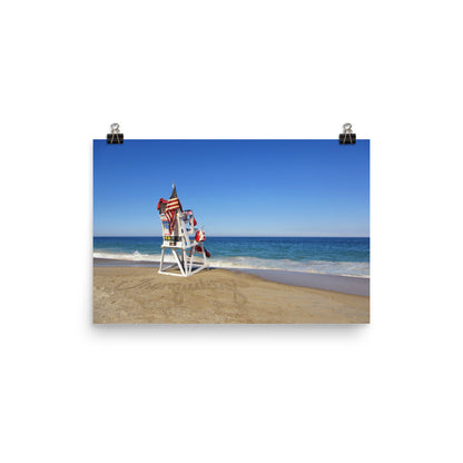 Image of Tranquility photographic art print on 12 inch by 18 inch premium luster photo paper by Jessica St. Clair depicting a lifeguard stand with an American flag overlooking calm ocean water with the word tranquility written in the sand